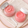 Open jars of pink sugar lip scrub and pink overnight lip mask with a small white applicator spatula on cream linen, top-down view