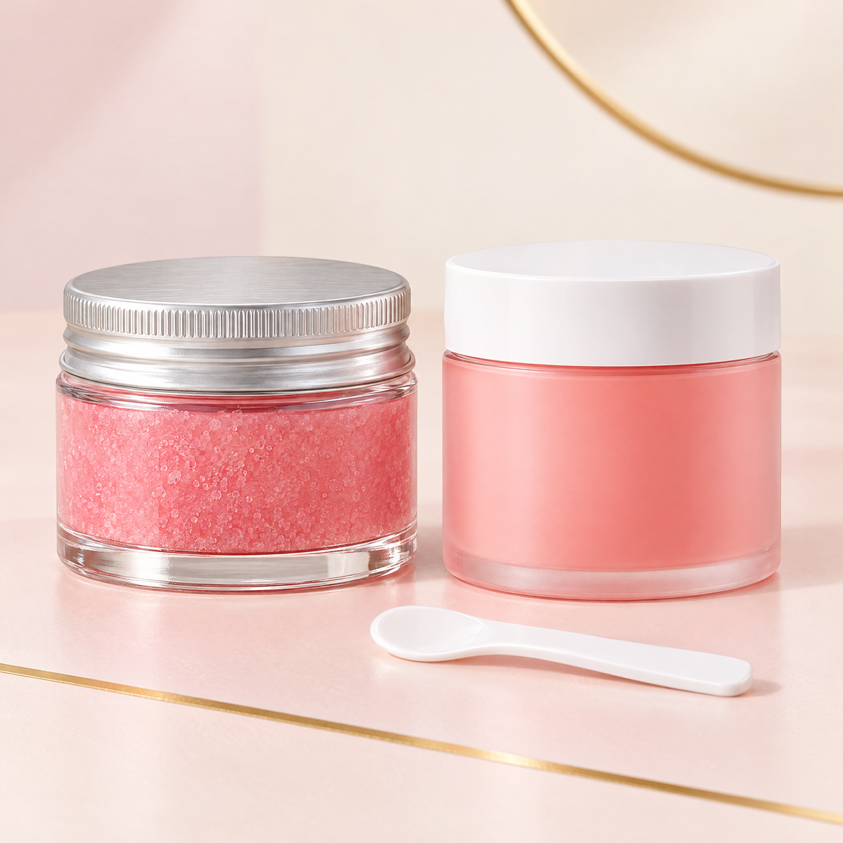Clear glass lip scrub jar with silver lid next to a coral pink lip mask jar with white lid and a white spatula on a blush backdrop